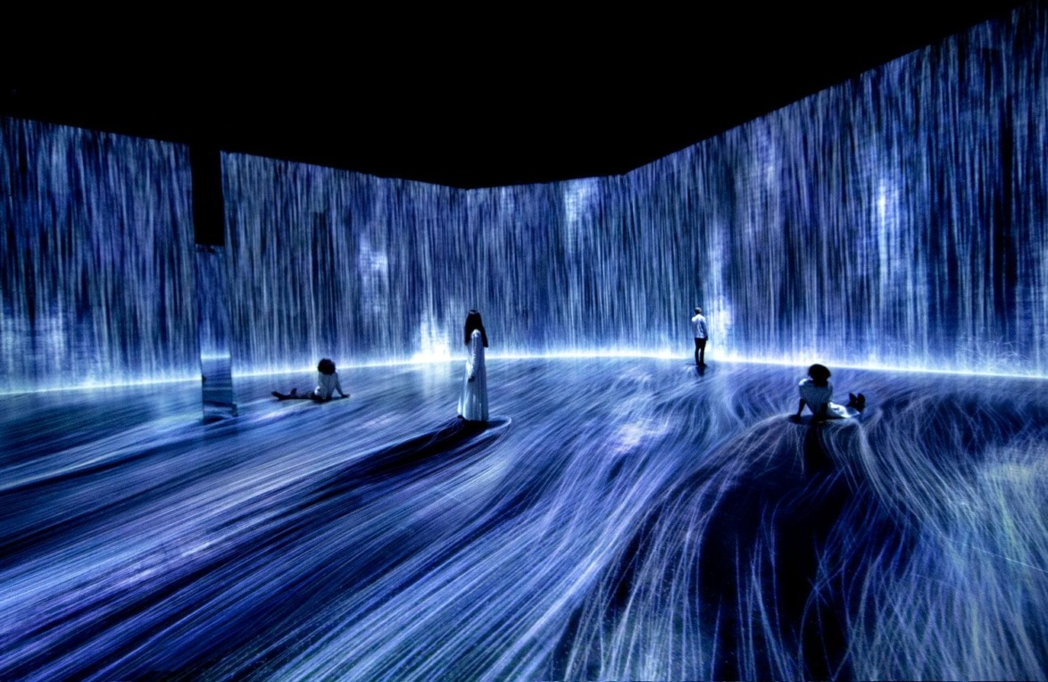 People interacting with digital waterfall art installation in a dark room with flowing light patterns on walls and floor.