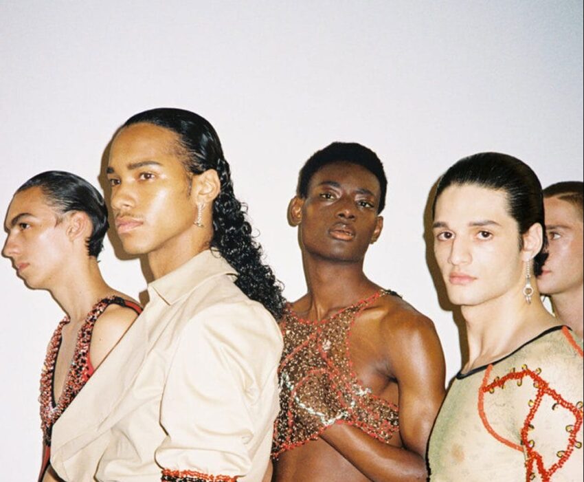 Group of four fashion models in stylish outfits, featuring unique hairstyles and colorful accessories, posing against a plain background.