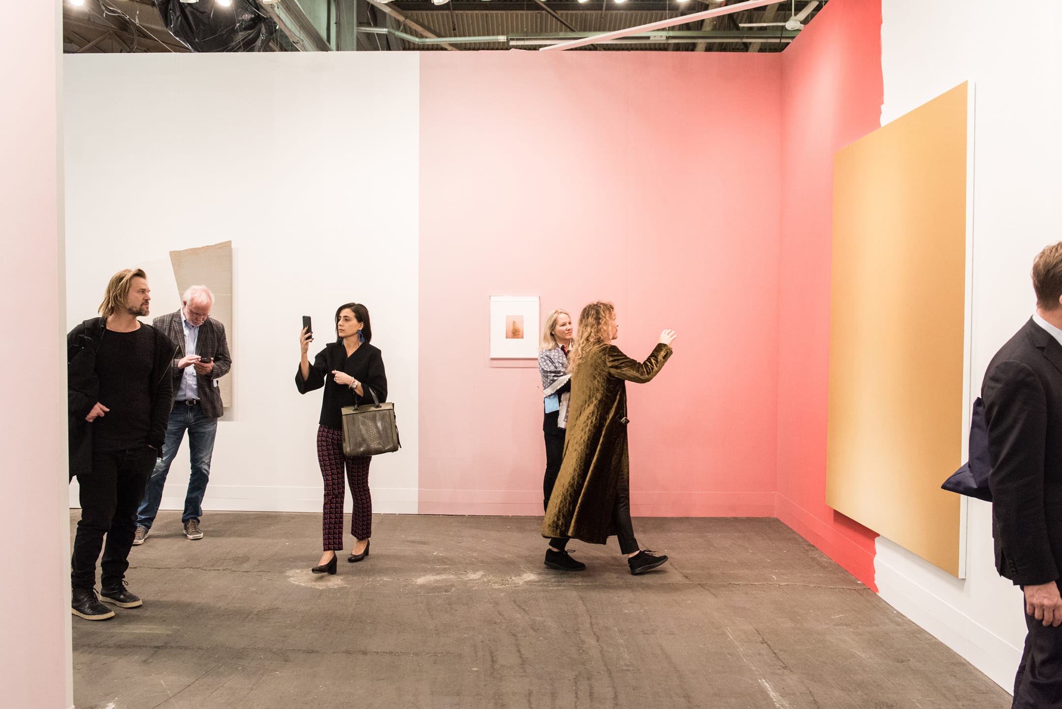 People viewing art pieces in a gallery setting with pink and white walls and a large yellow artwork displayed.