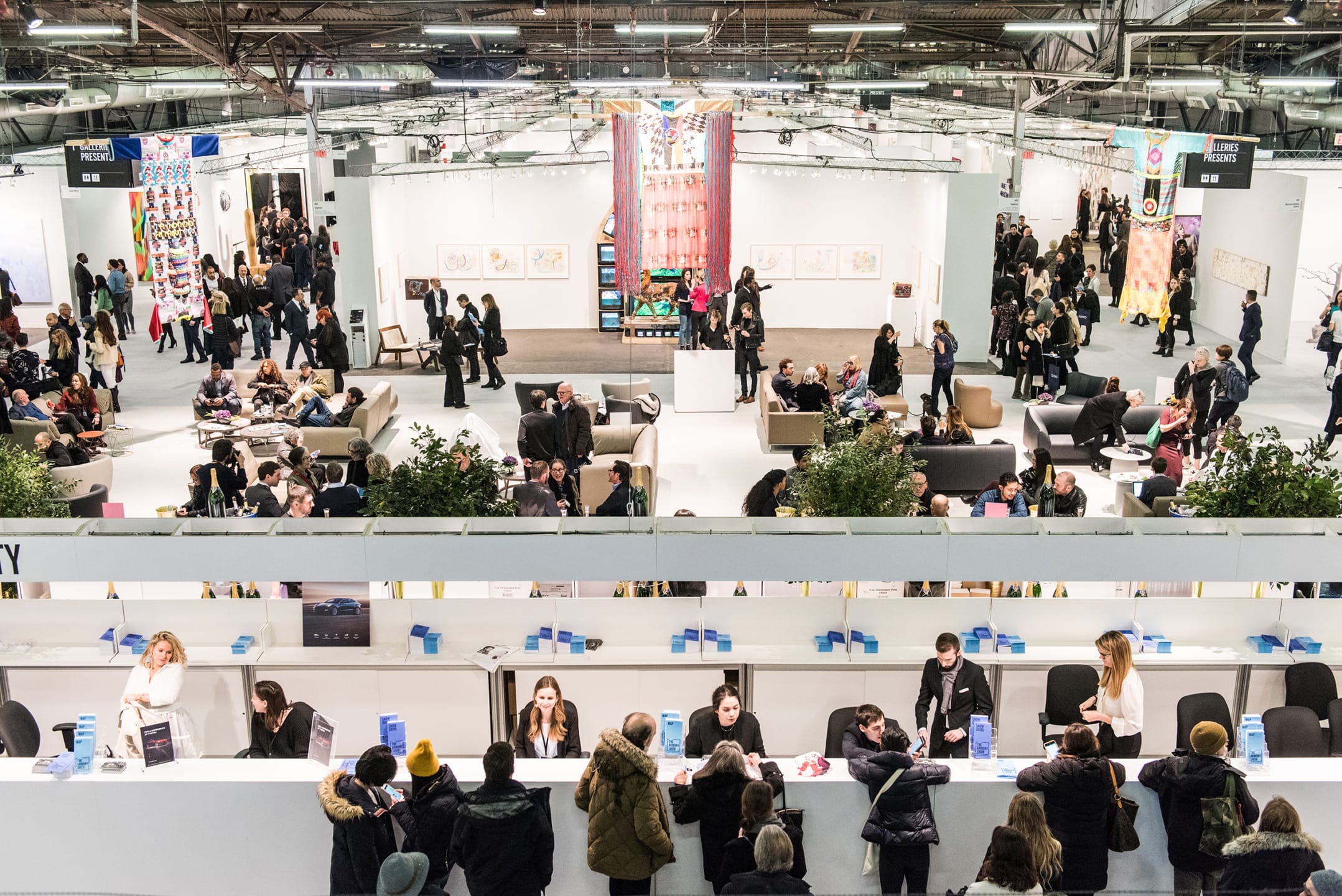 Art fair with attendees exploring exhibits, artwork displayed on walls, and people seated in conversation areas.