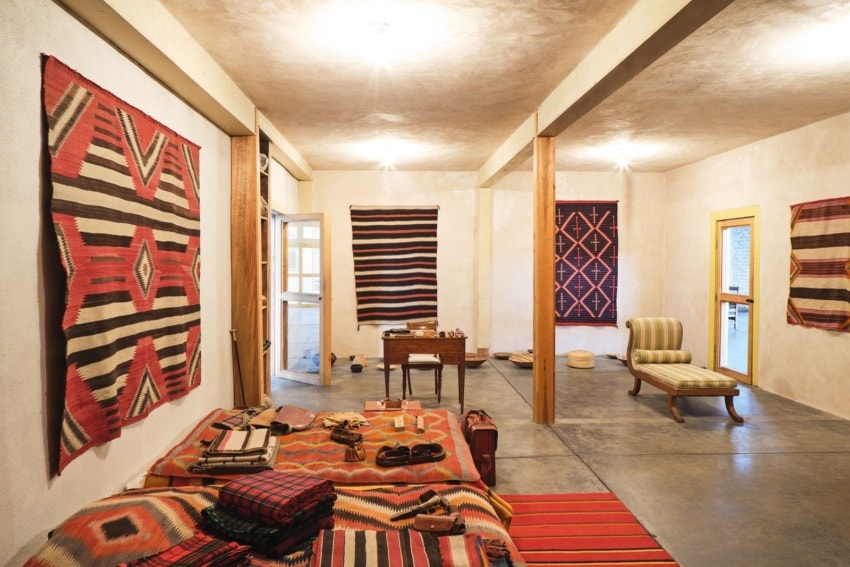 Room with colorful tribal rugs on walls and floor, wooden pillars, cushioned chair, and traditional items on a wooden table.