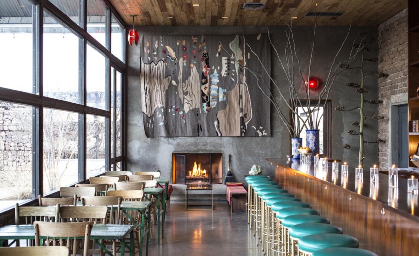 Cozy modern cafe interior with a fireplace, wooden ceiling, green bar stools, and large windows providing natural light.