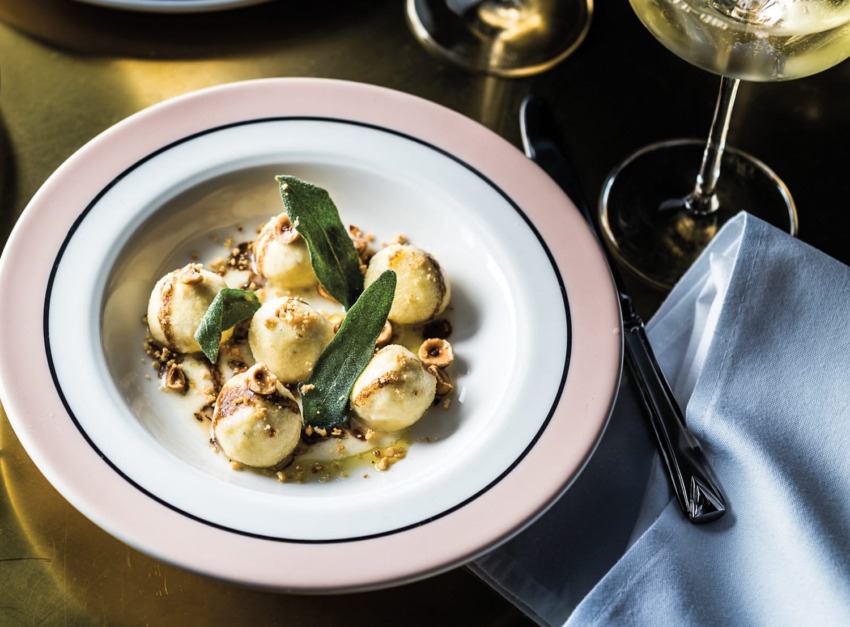Plate of ricotta-filled gnocchi with sage leaves and nuts on a table beside a glass of white wine.
