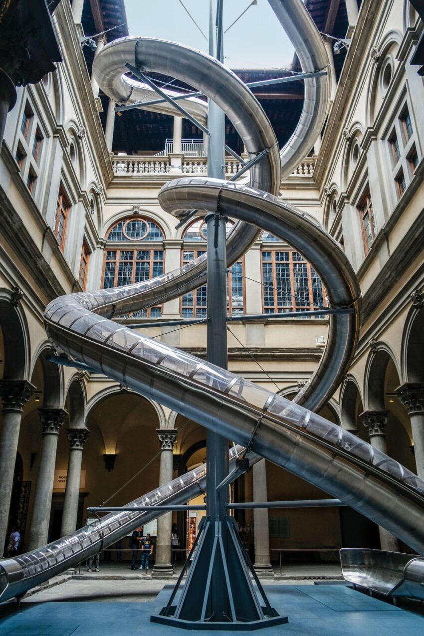 Large spiral slide art installation inside a historic building courtyard with arched windows and columns.