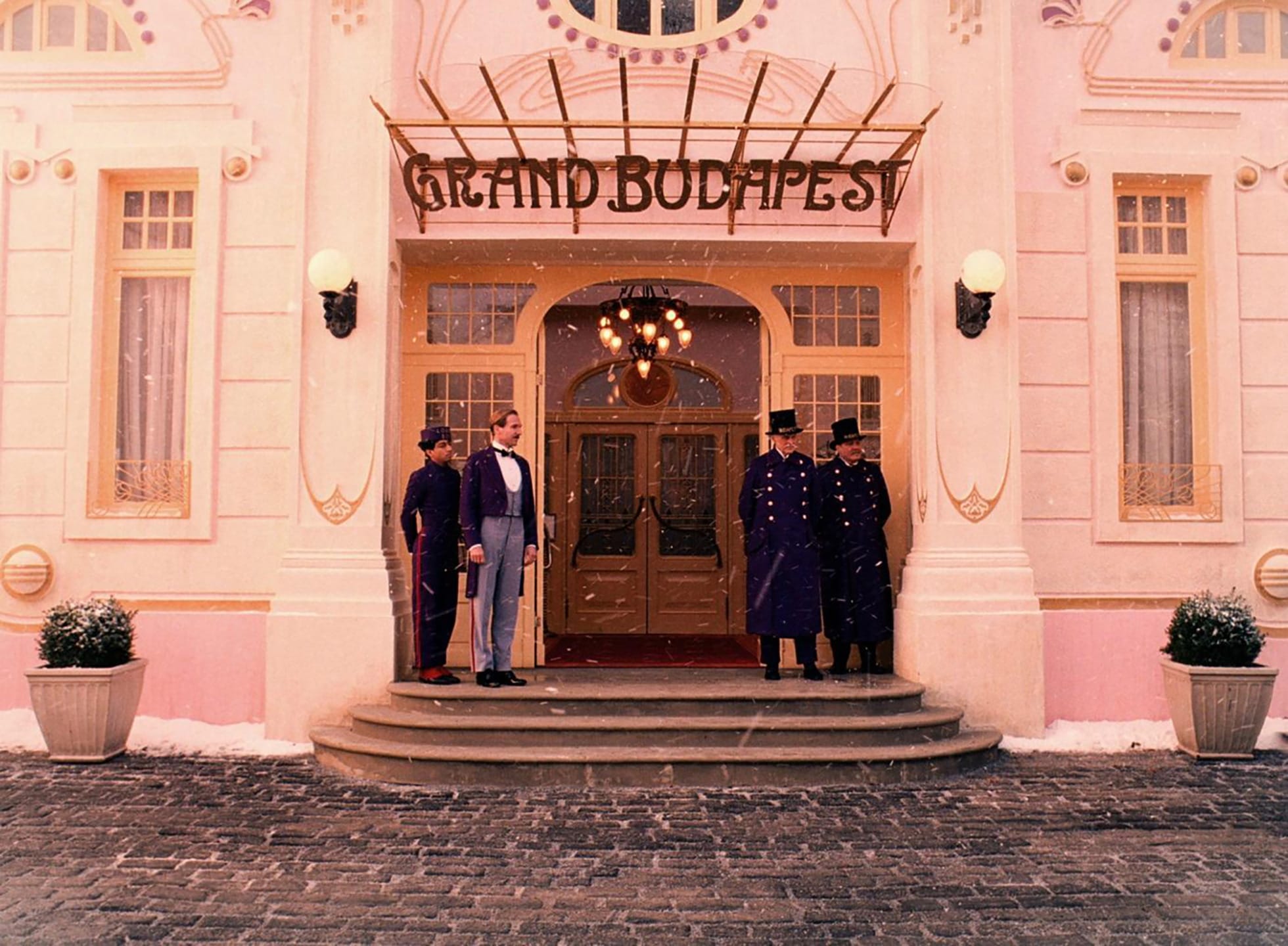 Front entrance of the Grand Budapest Hotel with staff and bellboys standing in uniform during a snowy day.