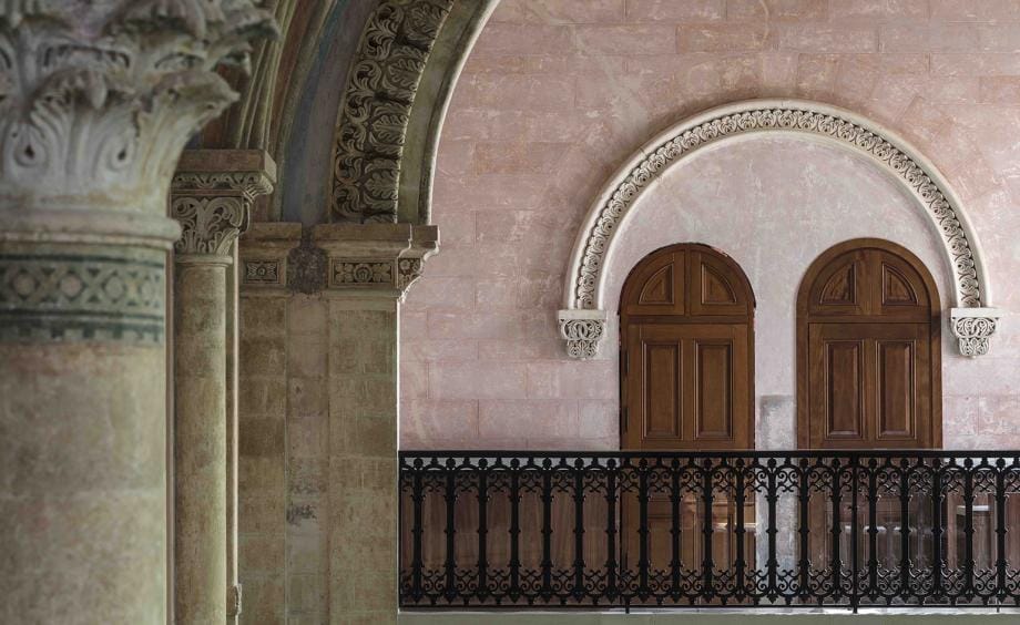 Ornate stone corridor with detailed arches, wooden doors, and wrought iron railing in a historic building.
