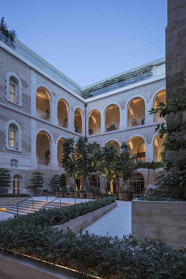 Courtyard with arches and greenery at twilight, featuring a multi-story building with illuminated windows and lush plants.