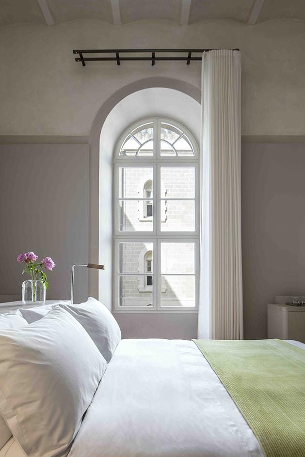 Elegant bedroom with arched window, green-accented bed, white curtains, and a vase of purple flowers on the bedside table.