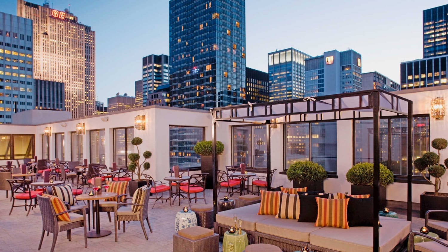 Rooftop terrace with modern furniture and city skyline view at dusk, including lanterns, plants, and stylish seating arrangements.