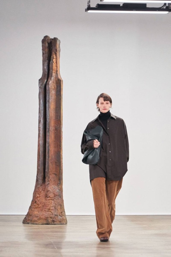 Person walking in fashion show wearing oversized clothing, carrying a bag, with large sculpture in the background.