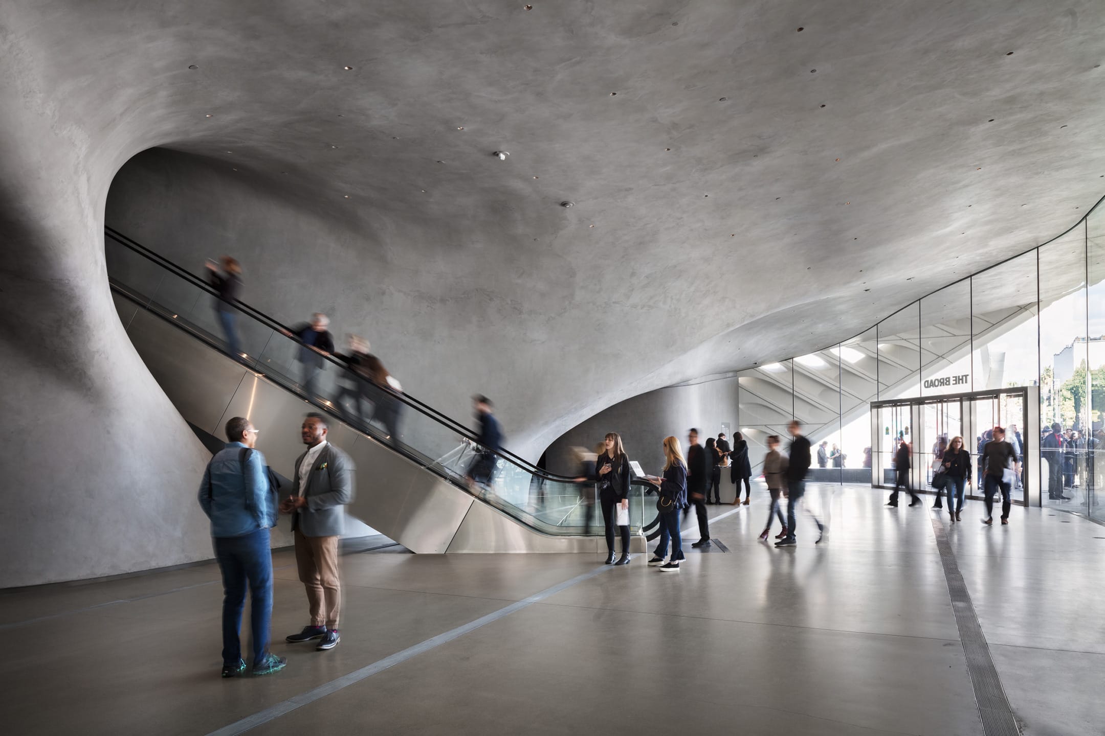 Museum lobby with modern architecture, people on escalator, and visitors walking or chatting, featuring curved concrete walls.