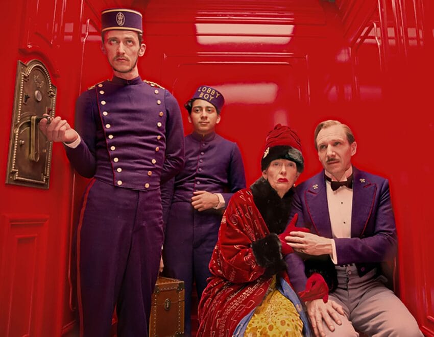 Hotel staff and guests in vintage attire sit and stand in a vibrant red elevator.