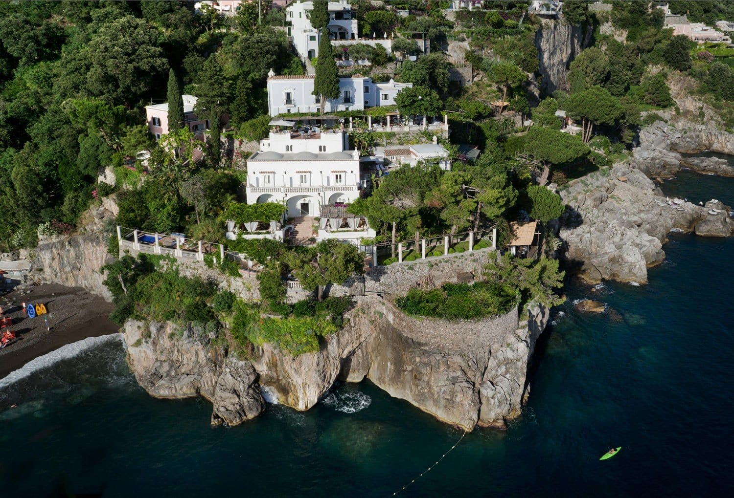 Aerial view of a villa perched on a cliff surrounded by lush greenery and overlooking the sea, with a small beach nearby.