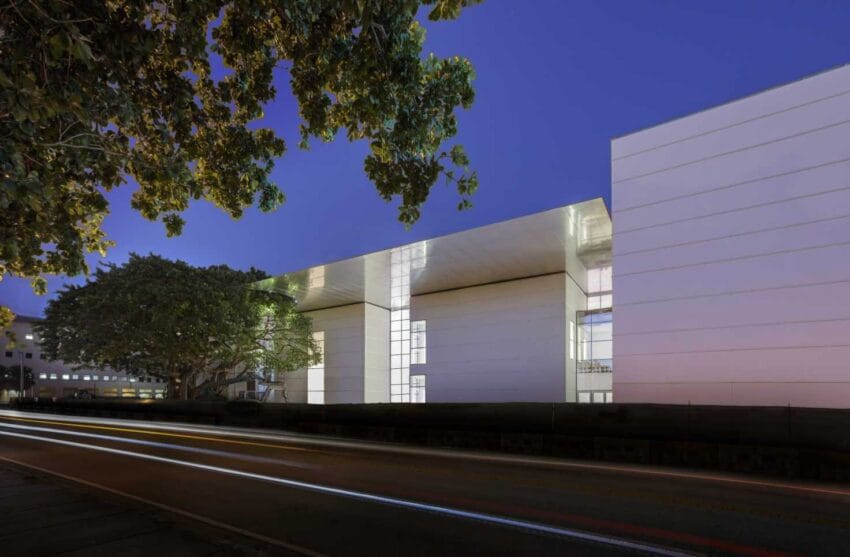 Modern white building with large windows illuminated at night, surrounded by trees, with a road in the foreground.