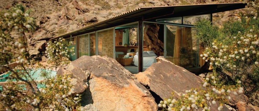 Modern glass house nestled among rocky desert landscape with blooming plants in the foreground.