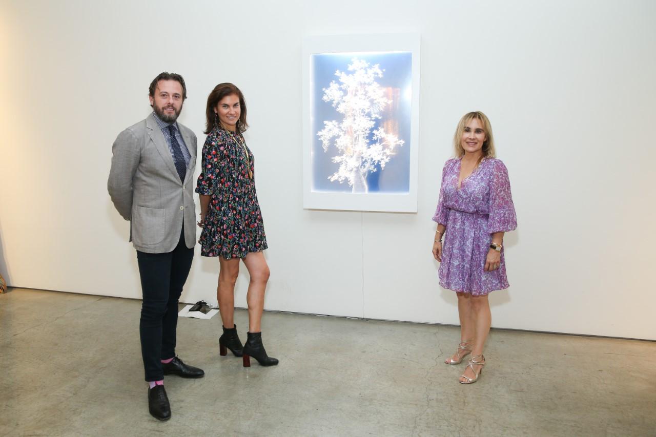 Three people standing in a gallery beside illuminated artwork on a white wall.