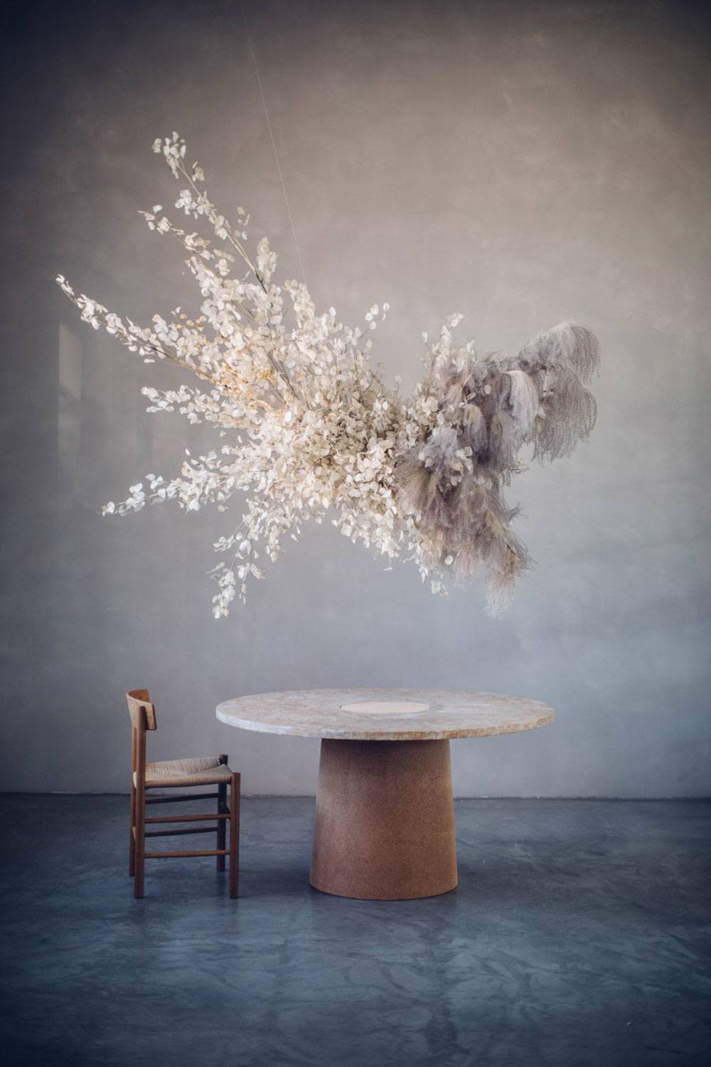 Minimalist room with round wooden table, single chair, and suspended floral arrangement against grey textured wall.