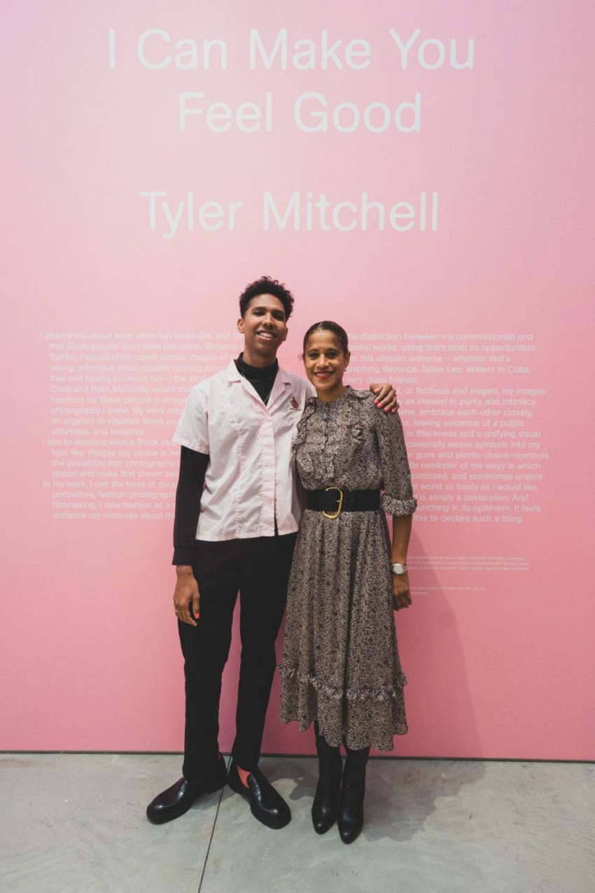 Two people standing in front of a pink wall with text, one in casual attire and the other in a patterned dress.