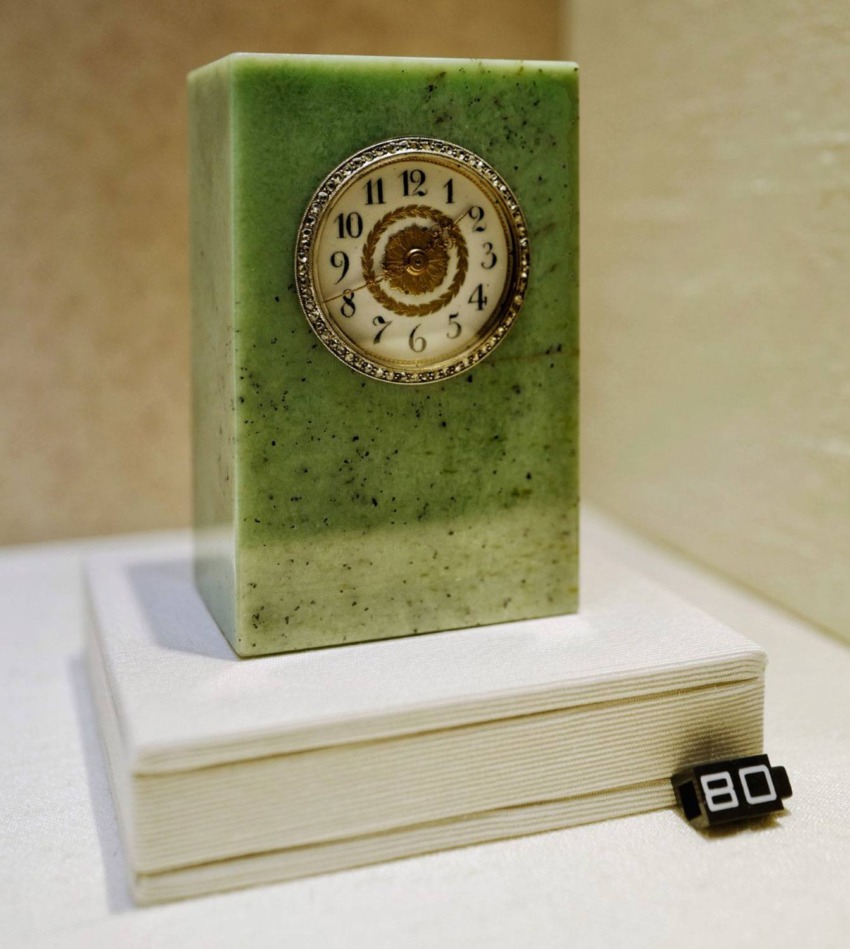 Green rectangular jade clock with ornate gold and white dial displayed on a stand with a numbered tag.