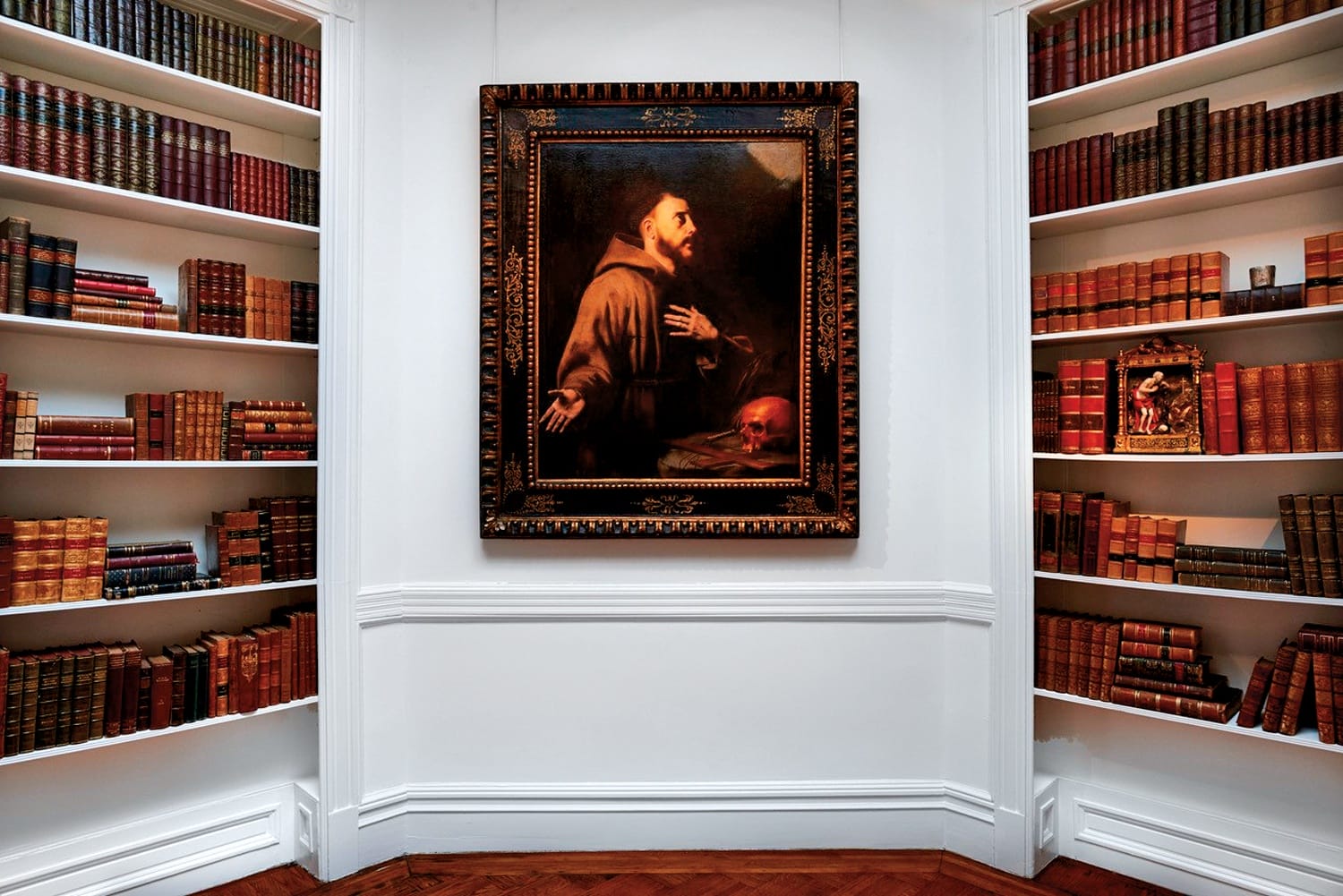 Painting of a monk in prayer hangs on a white wall, surrounded by shelves filled with books in a classic library setting.