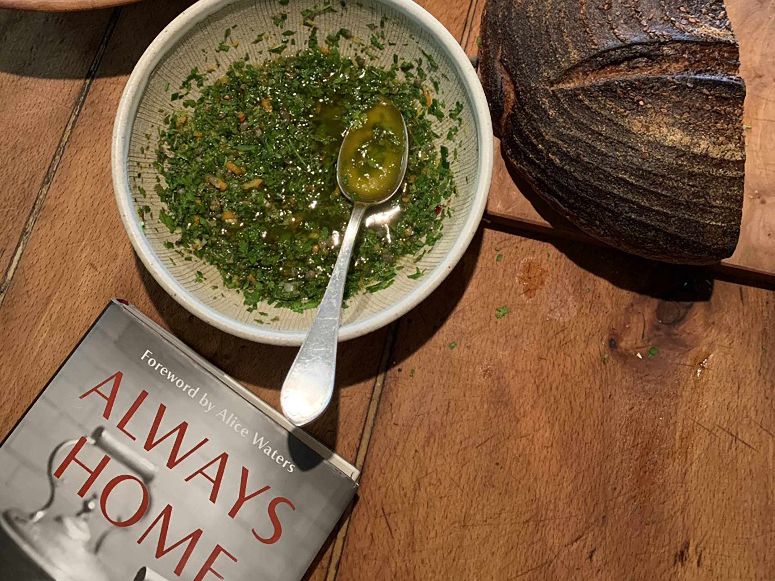 Bowl of herbs and olive oil mix with a spoon next to a book titled "Always Home" and a loaf of dark bread on a wooden surface.