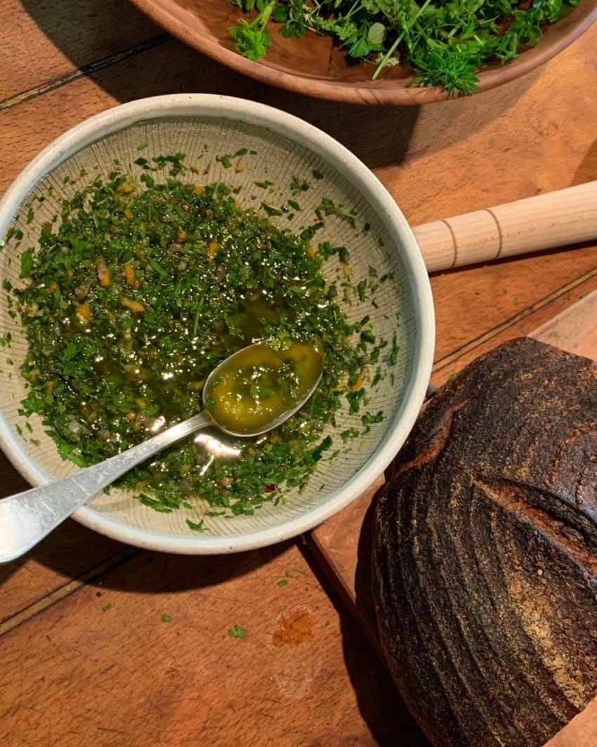 Herb-infused olive oil dip in a bowl with spoon, beside a crusty loaf of brown bread on a wooden table.