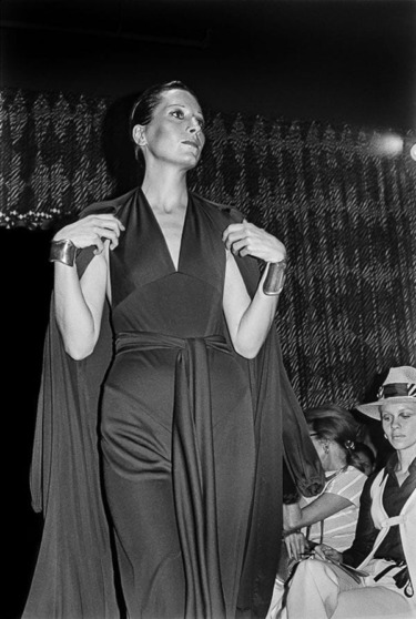 Model on runway wearing elegant black dress and large bracelets, audience in background watching fashion show