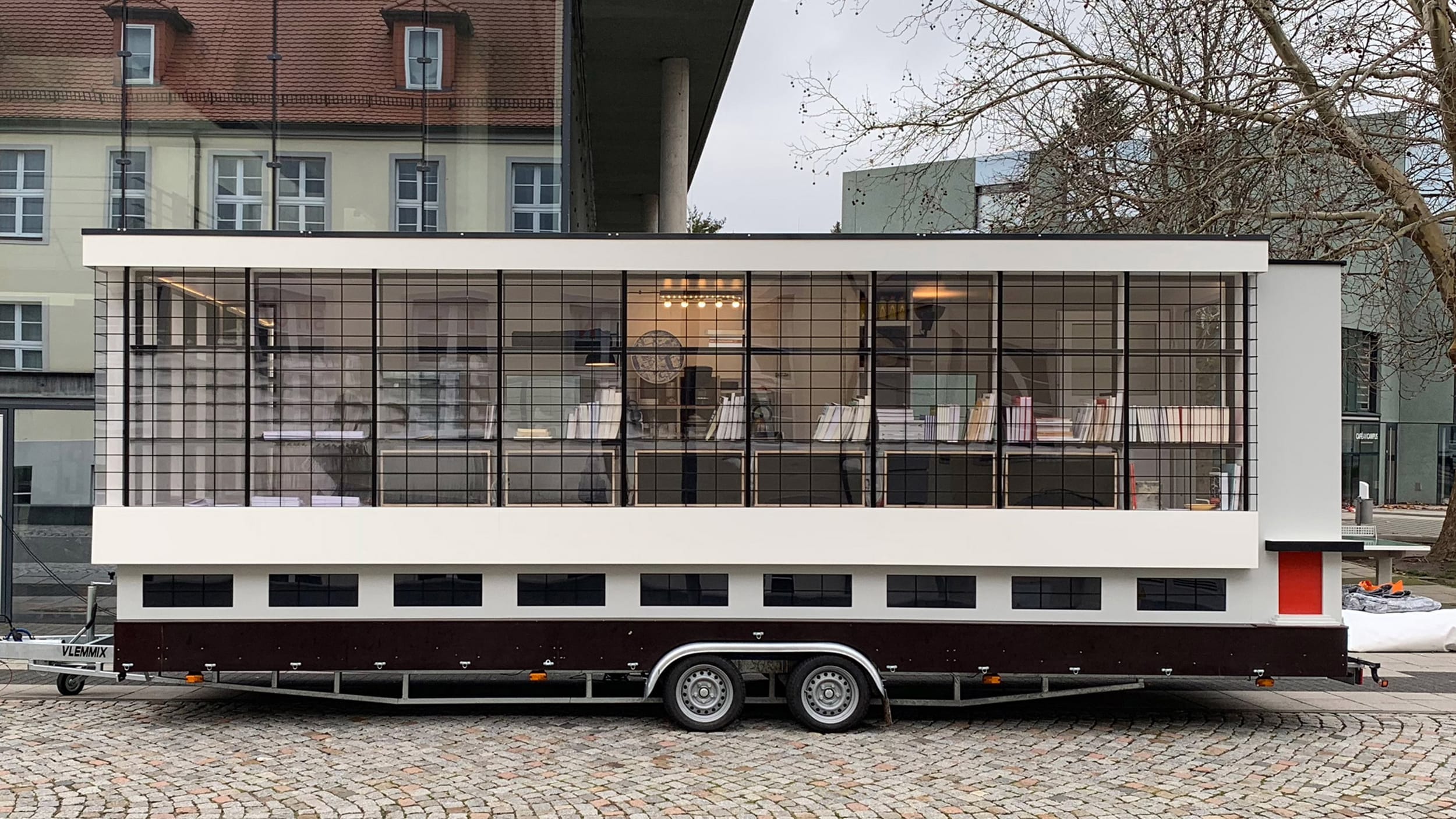 Mobile modern building on wheels with large windows displaying books and furniture, parked on a cobblestone street.
