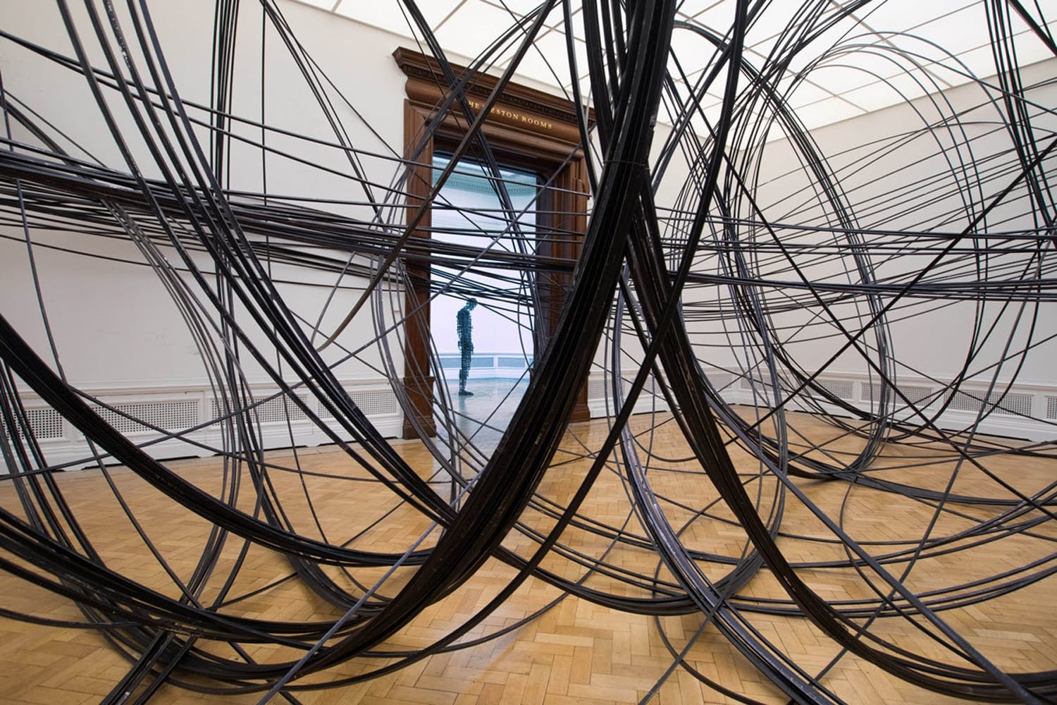 Room with large black cables crossing and intersecting, creating an intricate web-like installation on a wooden floor.