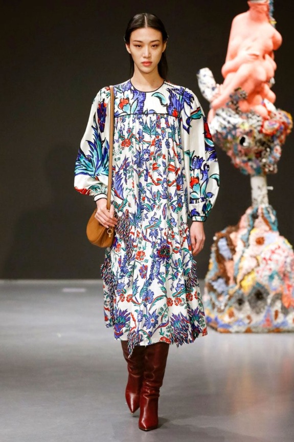 Model wearing a colorful floral dress and brown boots on a runway with an abstract sculpture in the background.