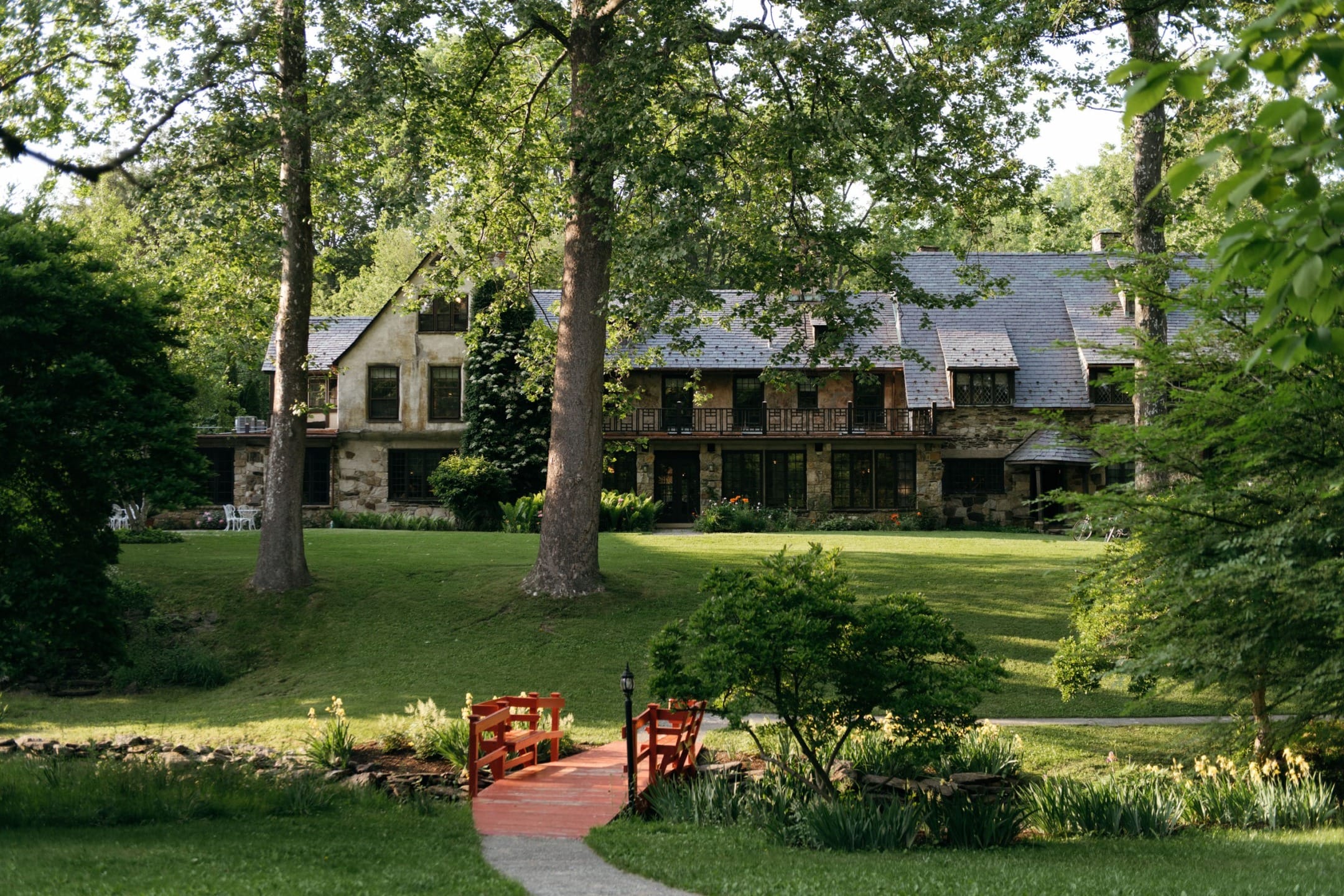 Large stone house surrounded by lush green trees and lawn with a red footbridge leading to the entrance.