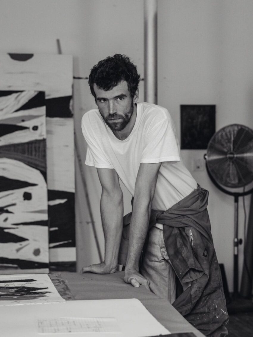 Man in a white t-shirt leaning on a table in an art studio with abstract paintings and a fan in the background.