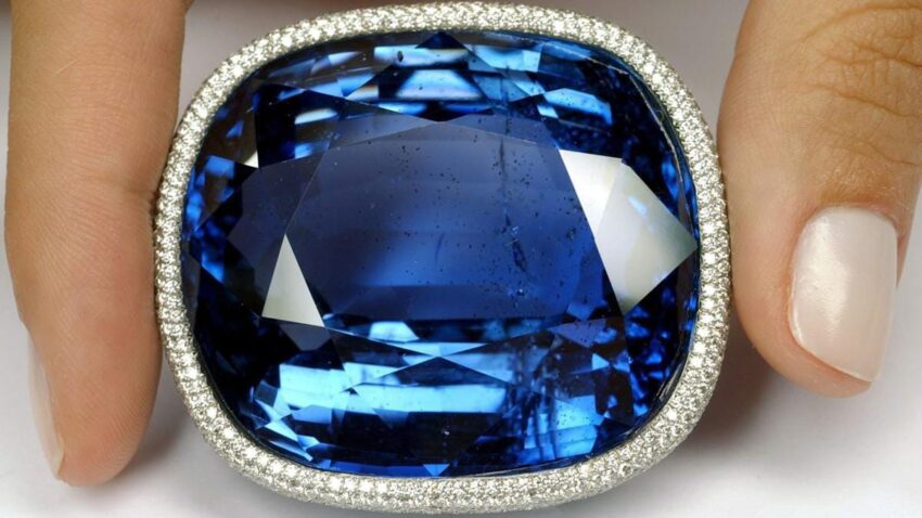 Large blue sapphire gemstone surrounded by small diamonds held by a hand.