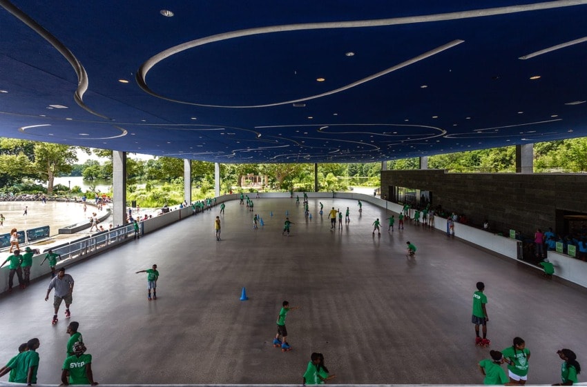 Children roller skating under a blue decorative ceiling in an indoor rink surrounded by trees on a sunny day.
