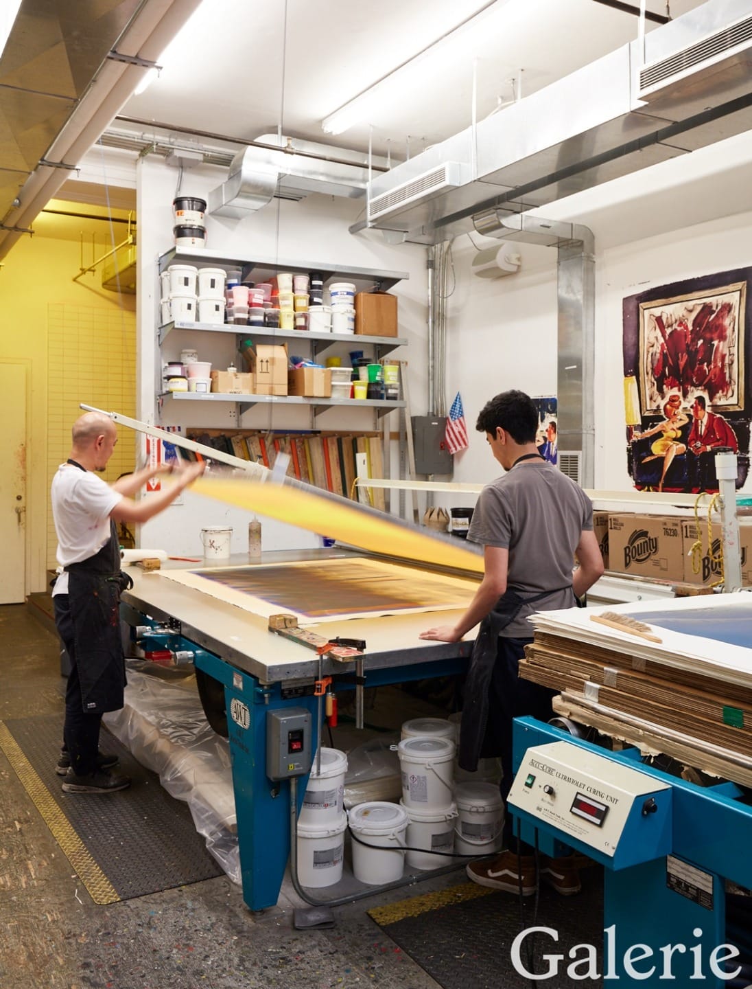 Two workers in an art studio preparing a canvas, surrounded by paint supplies and tools.