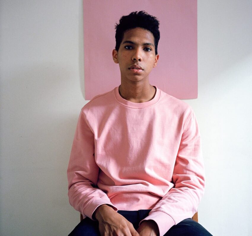 Person sitting on a chair wearing a pink sweater with a pink poster on the wall in the background.