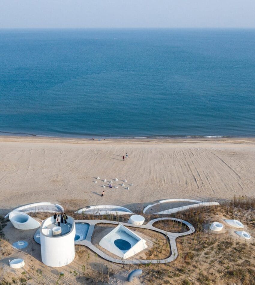 Aerial view of futuristic white beachfront building with circular and triangular shapes by a calm ocean.