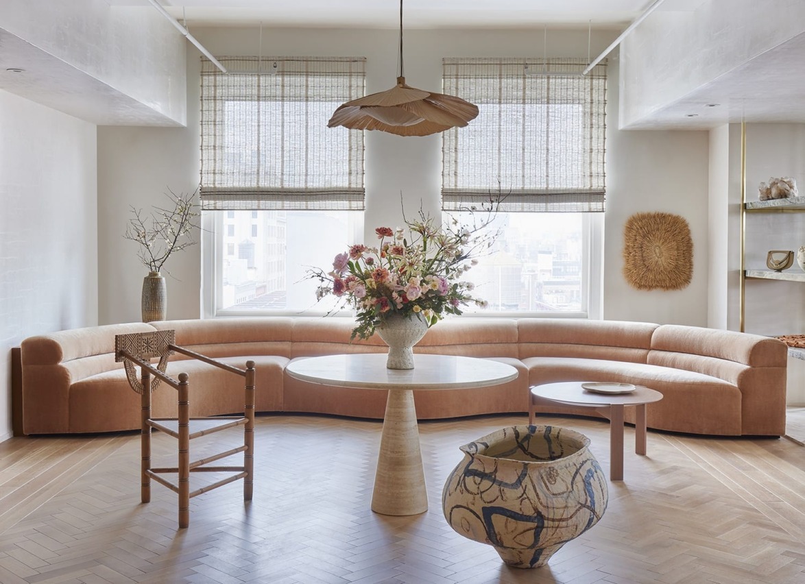Modern living room with a curved beige sofa, round table with flowers, artistic vase, and large windows with bamboo blinds.