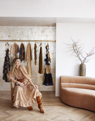 Woman sitting in a stylish room with hanging dresses, wearing a long patterned dress and brown boots.