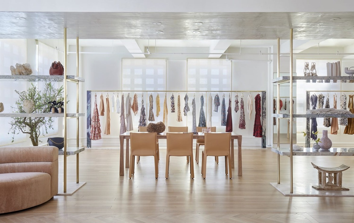 Elegant fashion showroom with hanging garments, a wooden table with chairs, and decorative shelves in a bright, modern space.