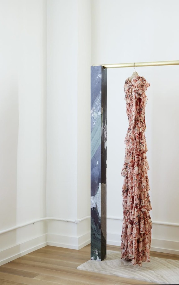Elegant floral ruffled gown hanging on a gold rail in a minimalist room with wooden floor and abstract art stand.