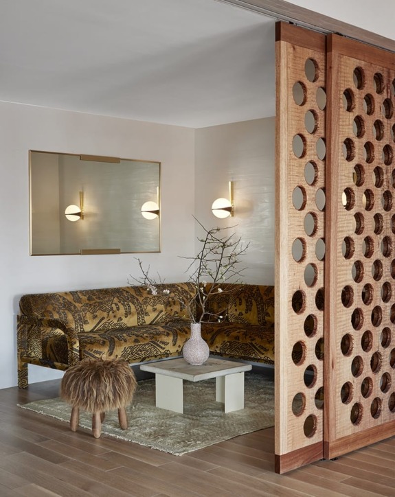 Modern living room with decorative wood divider, patterned sofa, wall sconces, round mirror, small coffee table, and fur ottoman.