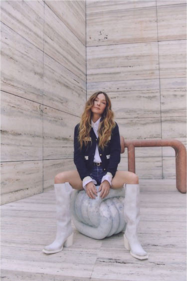Person sitting on marble sculpture wearing a navy blazer, white shirt, denim shorts, and white knee-high boots in a modern space.