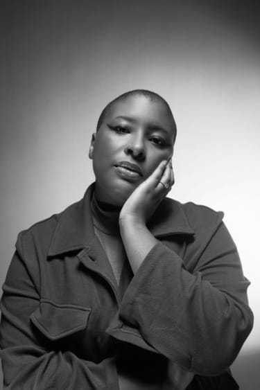 Person with short hair, wearing a jacket, posing thoughtfully with hand on cheek in a black and white photo.