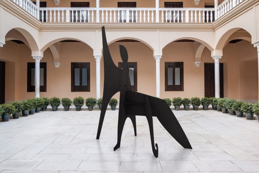 Abstract black metal sculpture in a courtyard with arched balconies and potted plants lining the perimeter.