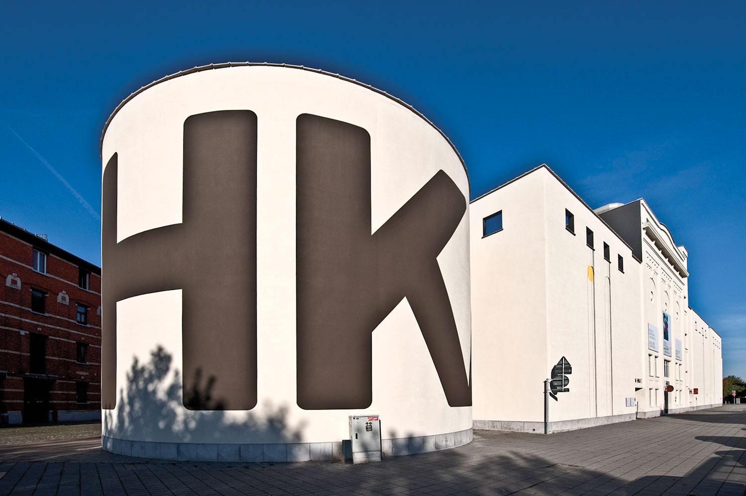 Modern cylindrical building with large black letters "HK" against a clear blue sky, adjacent to a larger white structure.