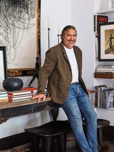 Man in a casual blazer and jeans sitting on a desk, surrounded by books and art in a stylish, modern room.