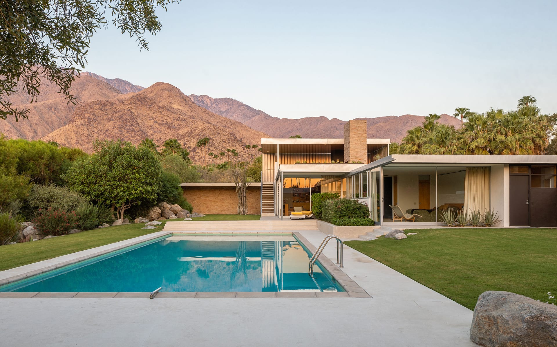 Modern house with large windows, pool, and mountain views in a landscaped yard.