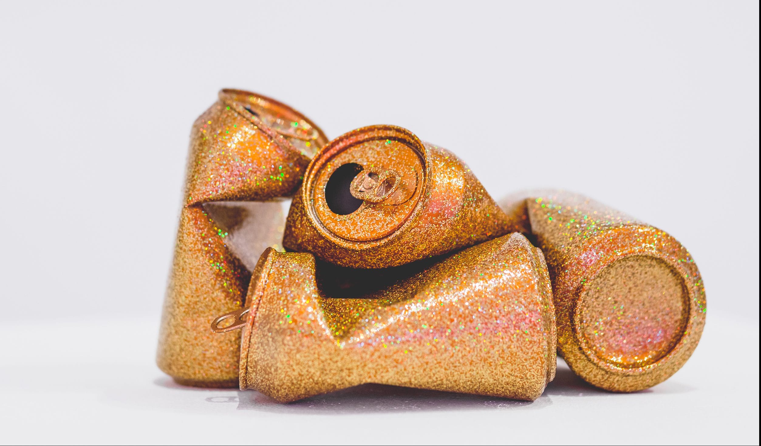 Crushed golden glittery soda cans stacked together on a white surface.