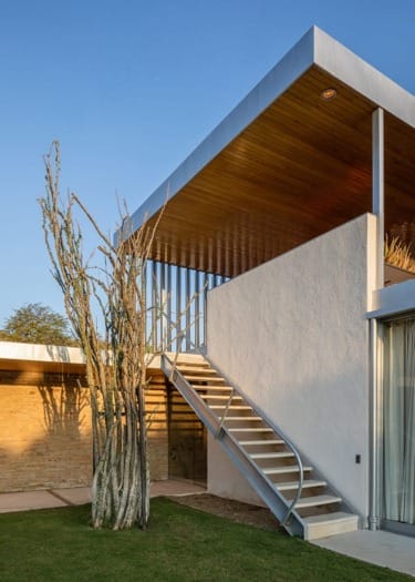 Modern house exterior with a metal staircase, wooden ceiling, and tall cactus plant under a clear blue sky.